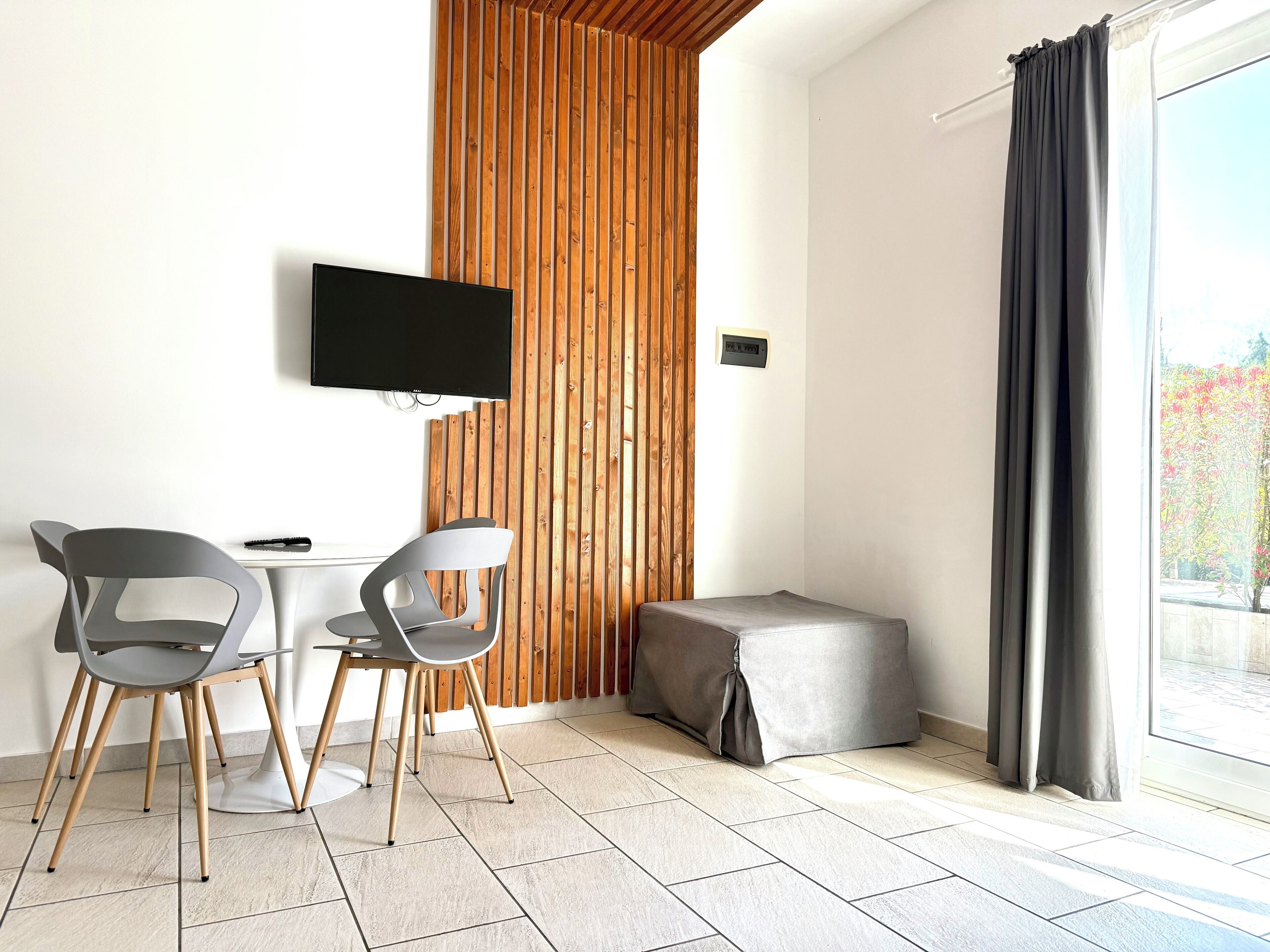 A modern room with a TV mounted on the wall, a wooden slat partition, a small table with two chairs, and a large window with curtains.