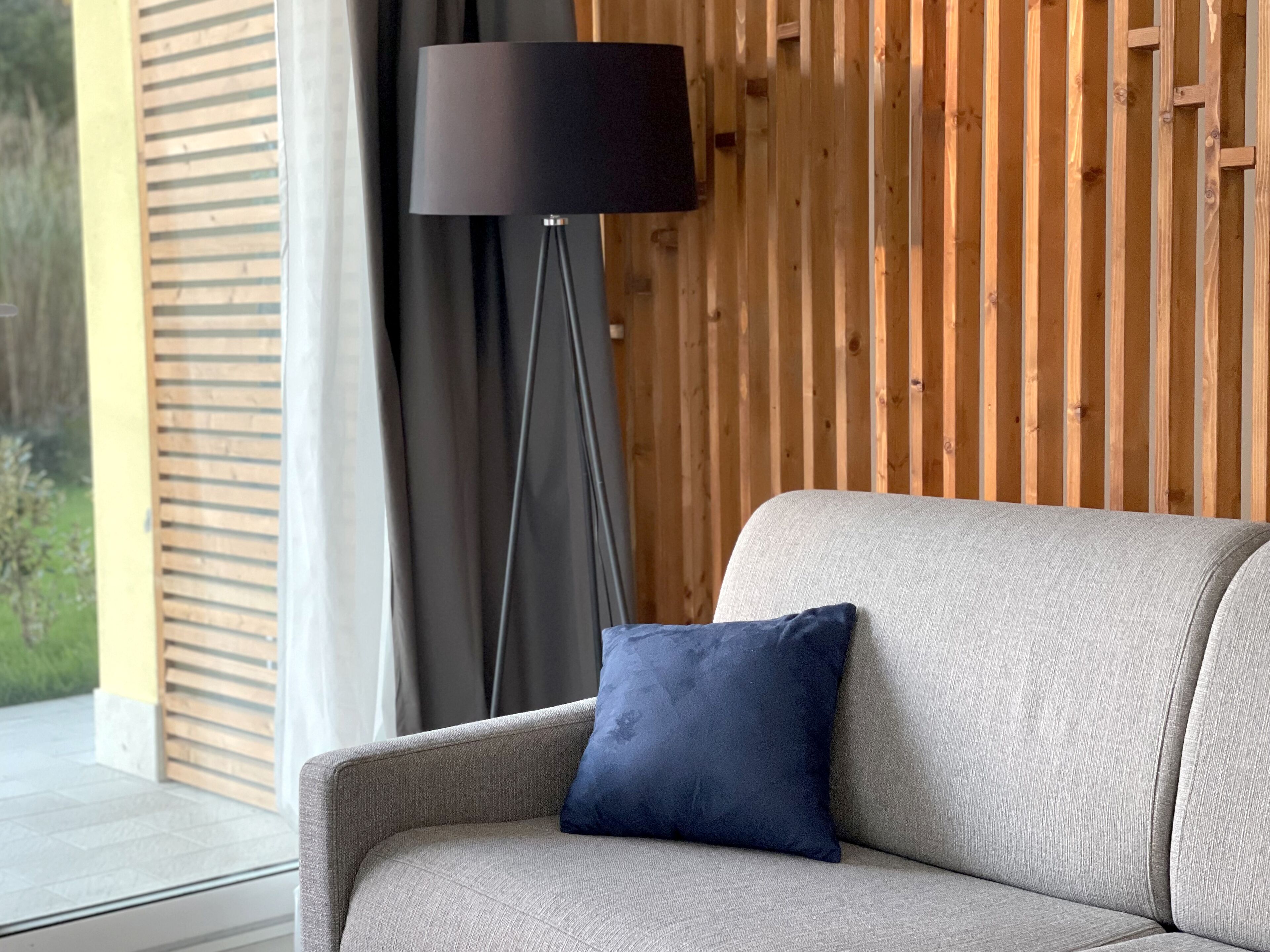 Ein modernes Wohnzimmer mit einem grauen Sofa, einem dunkelblauen Kissen und einer Stehlampe an einer Holzwand.