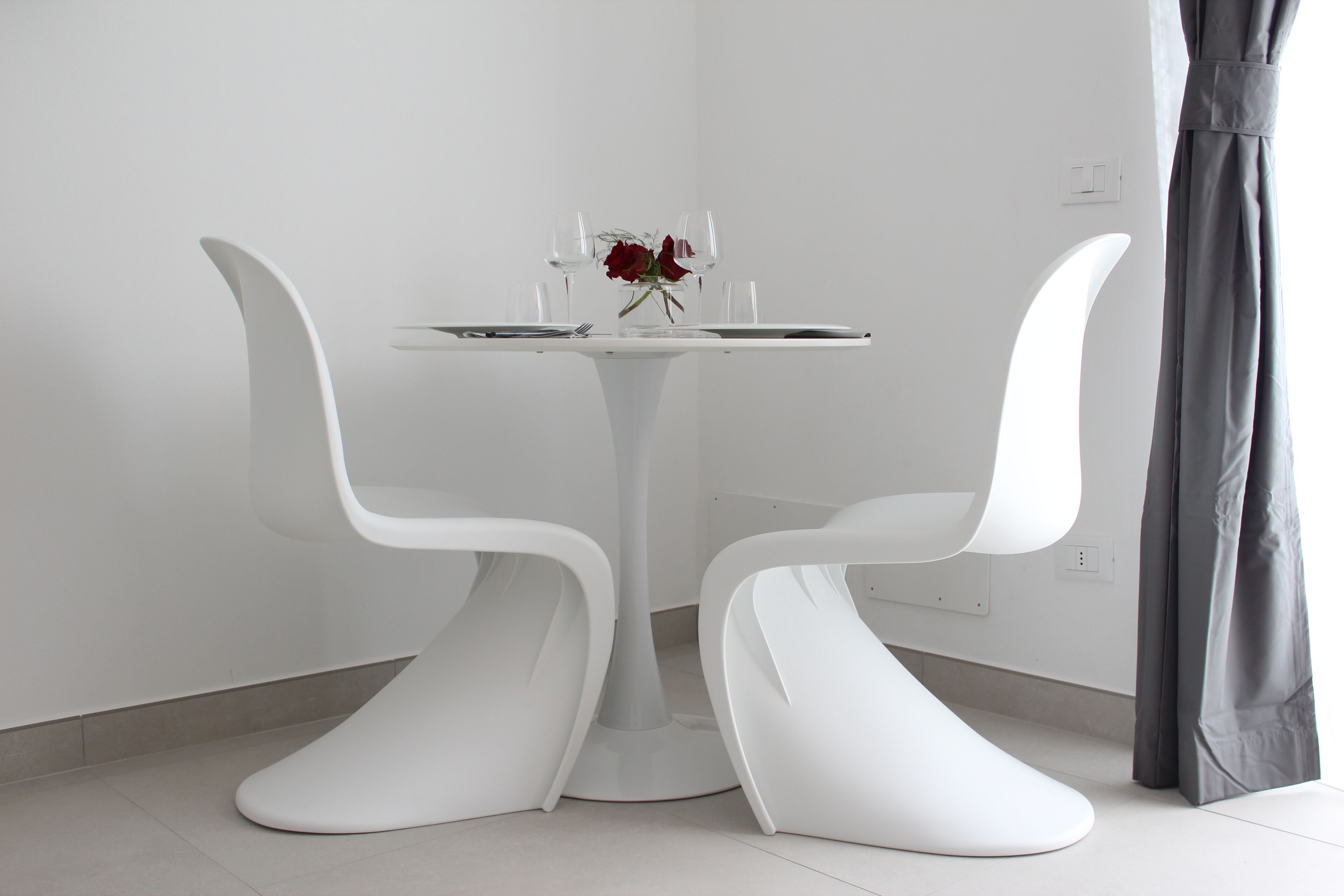 A modern dining area with a white table, two white chairs, a vase with a red flower, and a bright pendant light.