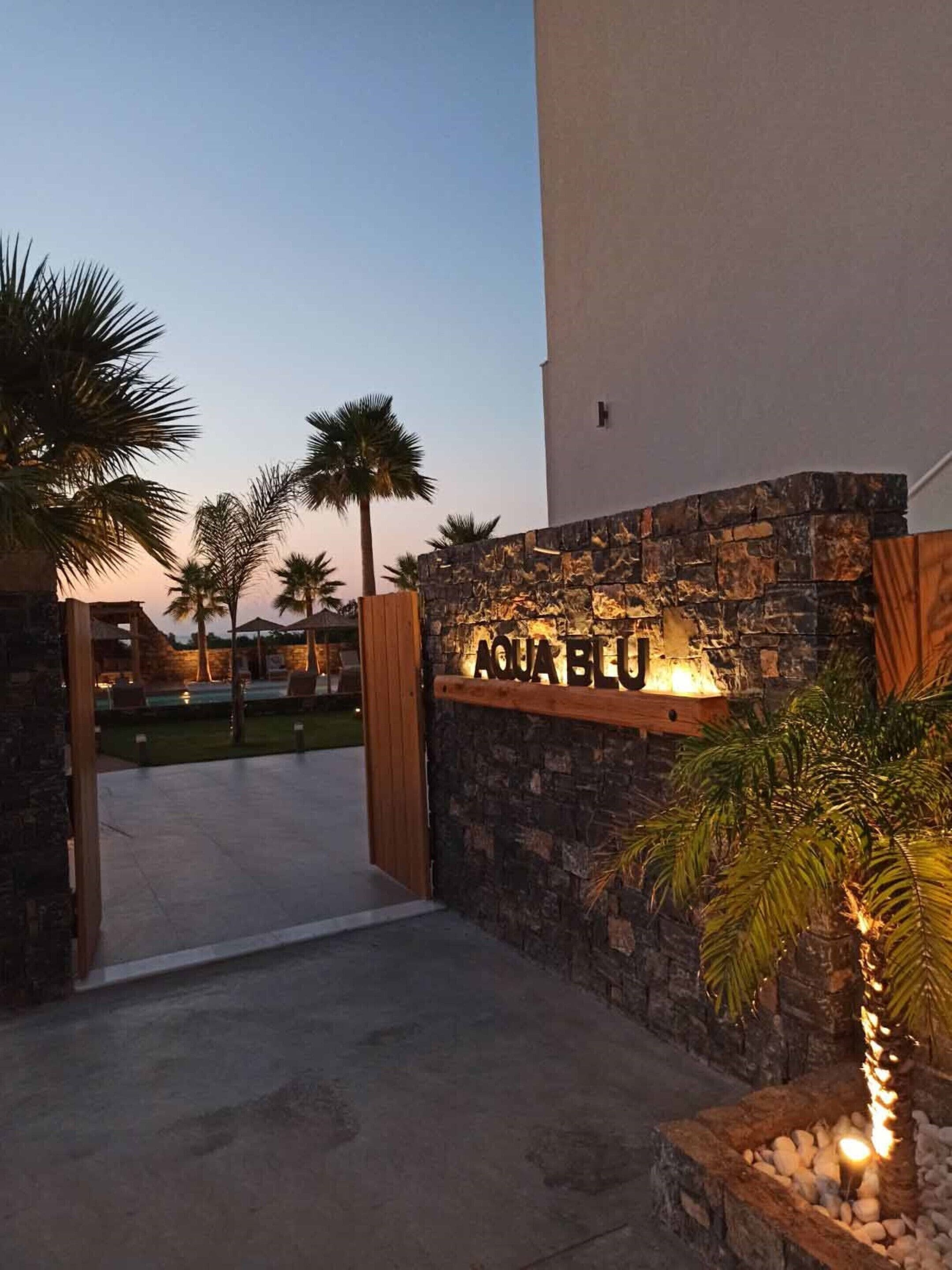 Een stenen muur met het verlichte bord 'AQUA BLU', palmbomen en een heldere lucht.