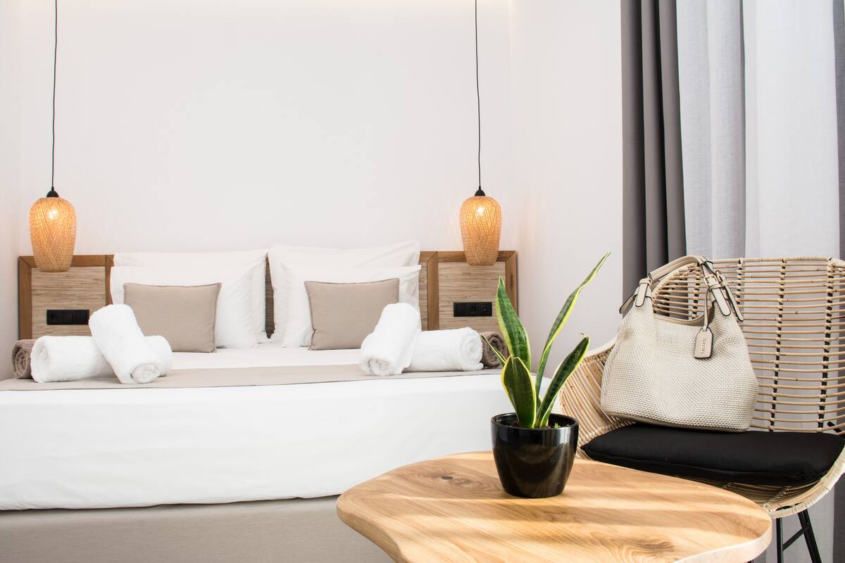 Une chambre d’hôtel bien rangée, avec un lit, deux lampes suspendues, une plante en pot, un fauteuil en osier et une petite table en bois.