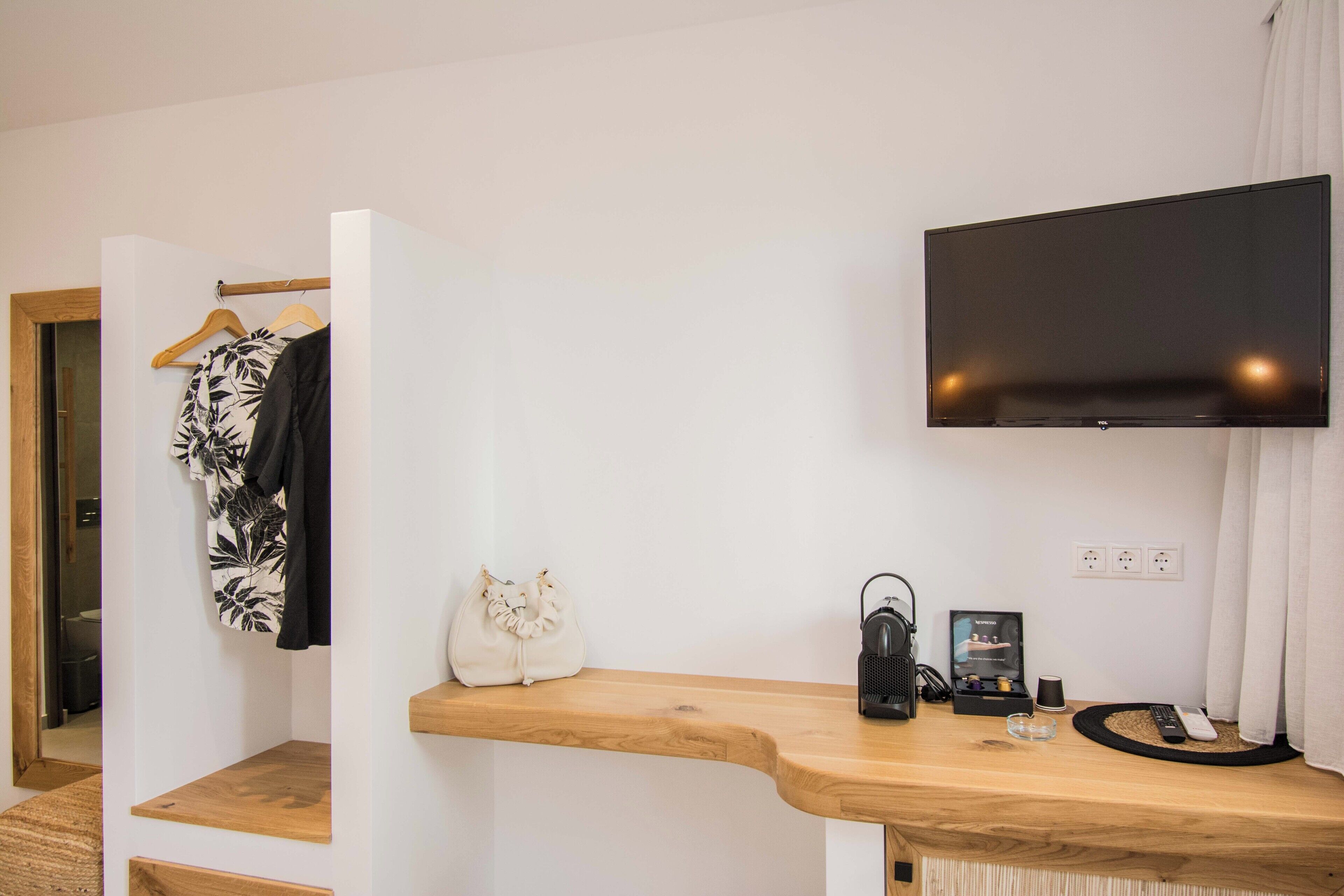 Une chambre d’hôtel avec une console en bois, un téléviseur à écran plat fixé au mur, un manteau suspendu à un porte-manteau et une petite table de chevet avec une lampe et des télécommandes.