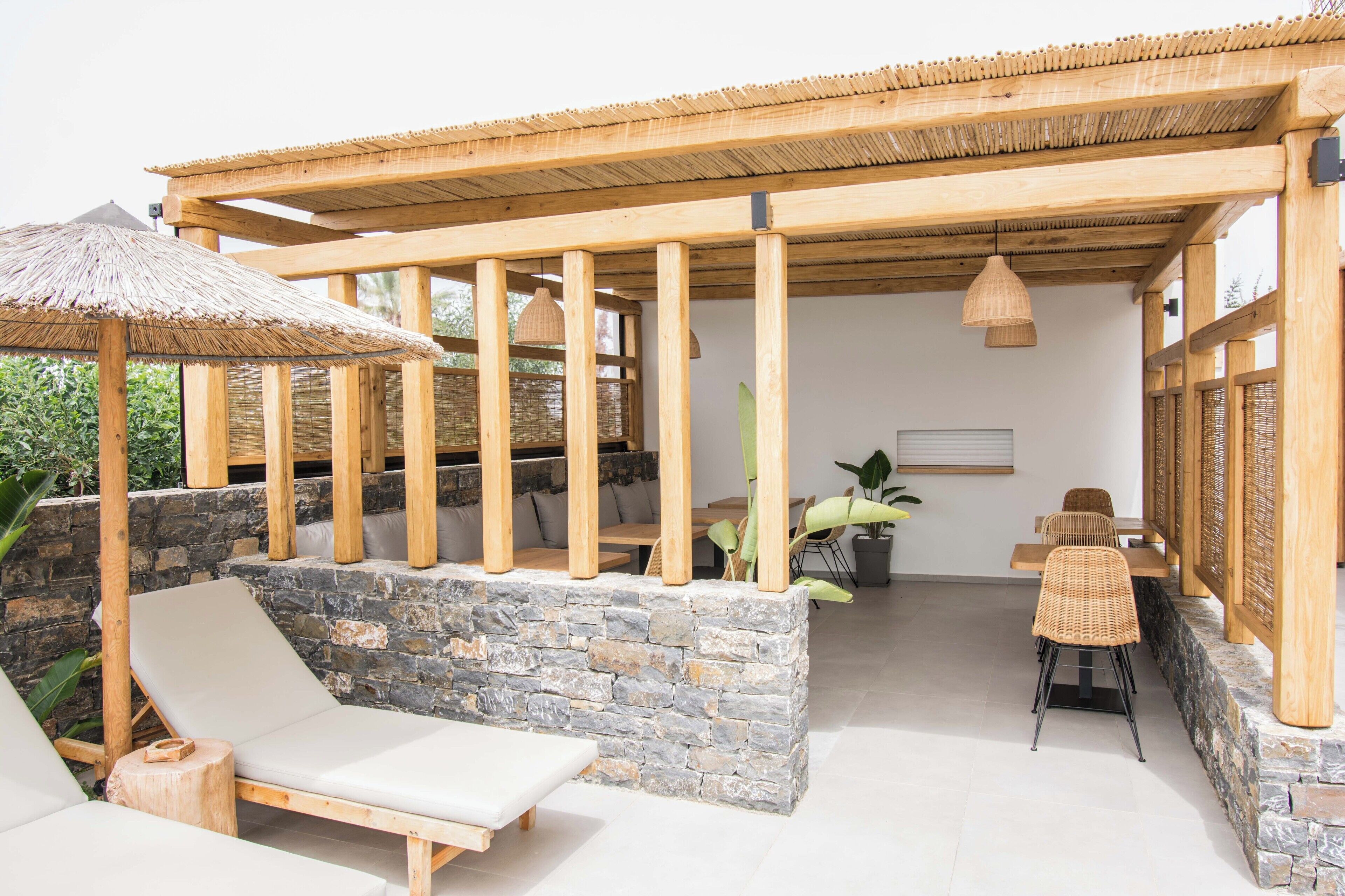 Un área de estar al aire libre con techo, paredes de piedra, mobiliario de madera y un paraguas de paja.