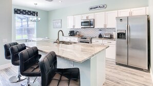A modern kitchen with white cabinets, stainless steel appliances, a marble island, and black bar stools. - 5BR Disney Villa: Pool & Spa, Game Room, Theater (Kissimmee)