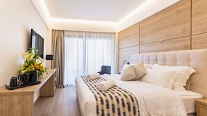 A modern hotel room with a large bed, a wooden headboard, a bedside table with a lamp, and a view of the outdoors through sheer curtains. - Hotel Acandia (Rhodes)