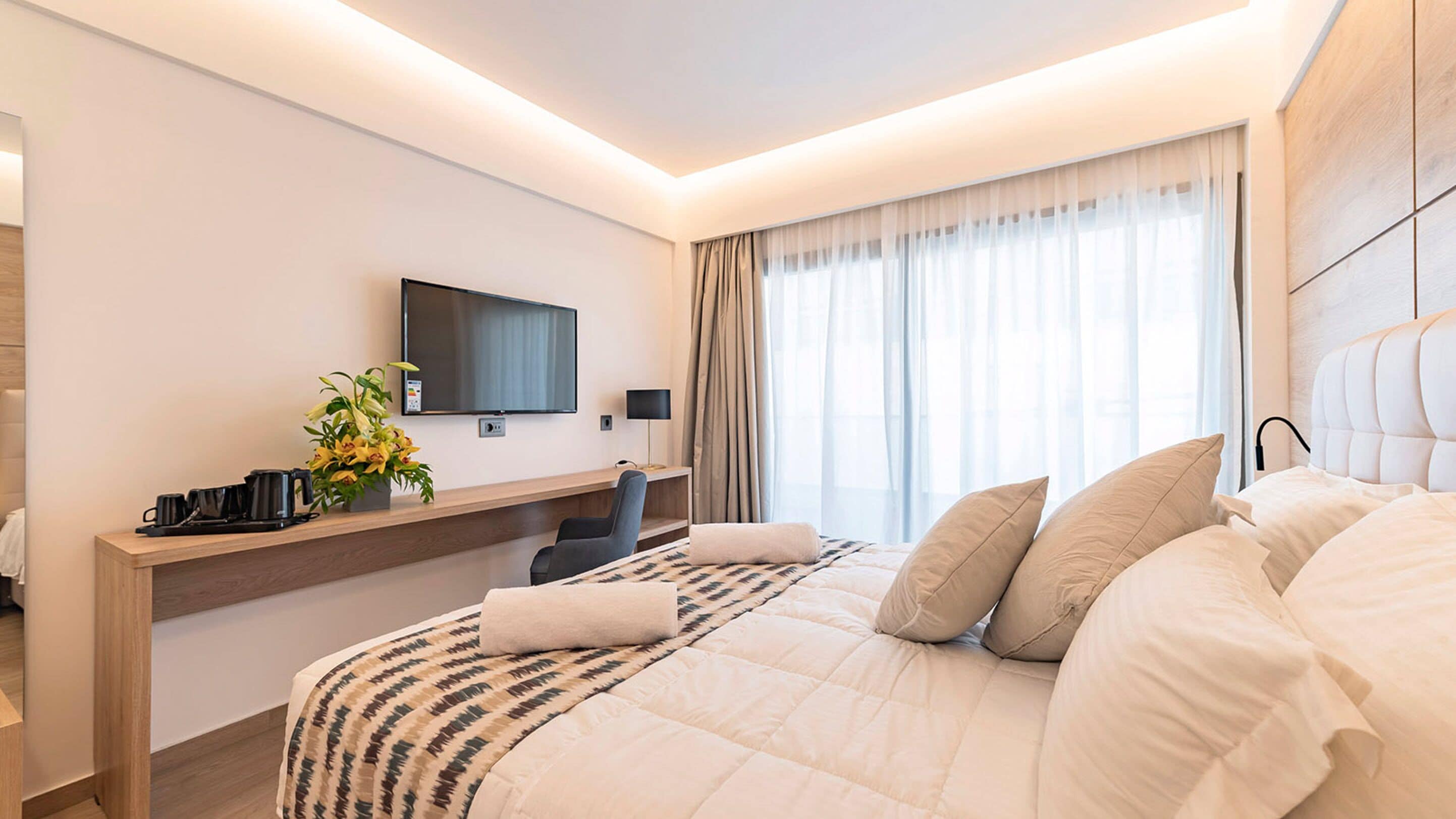 A modern hotel room with a large bed, a desk with a chair, a flat-screen TV, and a window with curtains.