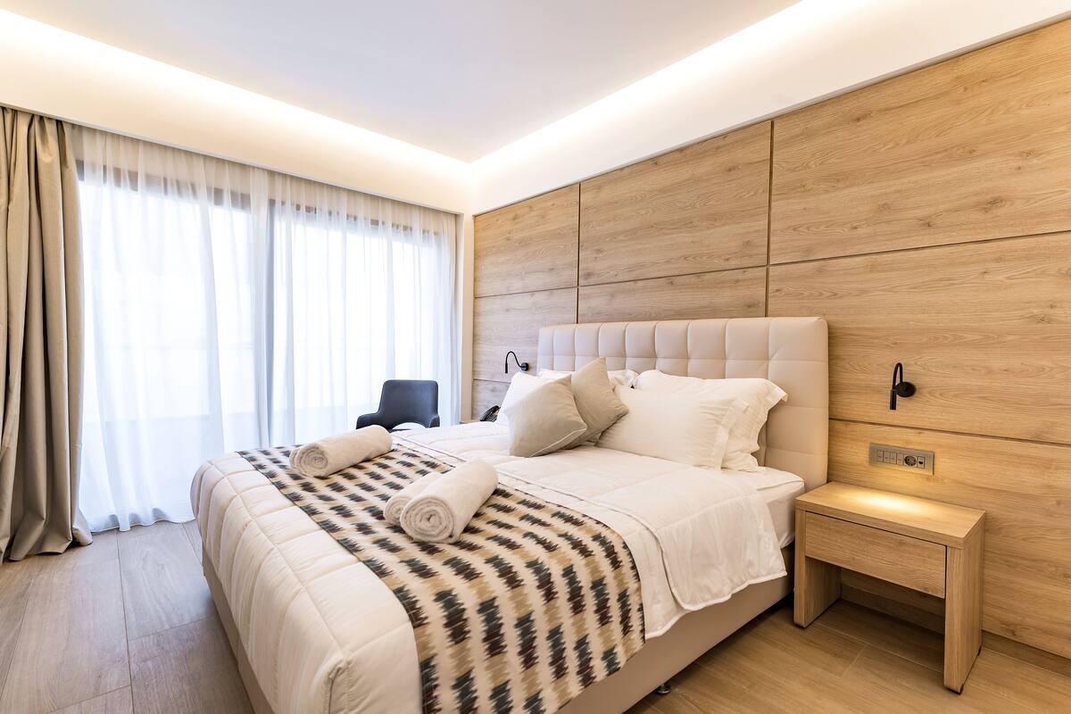 A modern hotel room with a large bed, a bedside table, a chair, and a wooden headboard.