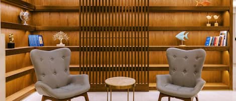 Une pièce comprenant deux fauteuils gris à revêtement en tissu, une petite table ronde, des étagères en bois avec des livres et des objets décoratifs, et une cloison en bois à lattes.