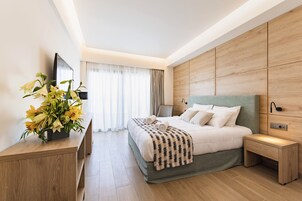 Quarto de hotel moderno com uma cama grande, cabeceira de madeira, mesa de cabeceira, televisão sobre um suporte e uma janela com cortinas.