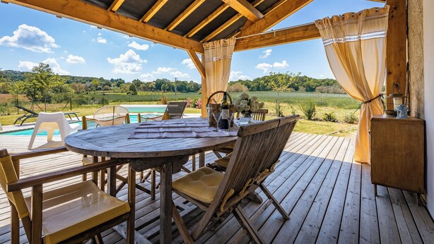 An outdoor wooden deck with a round table and chairs, overlooking a swimming pool and a scenic landscape. - holiday home with private pool and large covered terrace. (La Bachellerie)