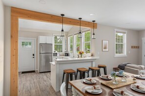 Une cuisine moderne dotée d’un îlot central, de poutres en bois, de luminaires suspendus et d’un coin repas avec une table en bois prévue pour quatre personnes.