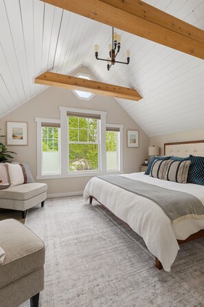 Une chambre avec un plafond en pente, un grand lit, une table de chevet, une chaise et une fenêtre donnant sur des arbres.