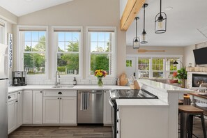 A modern kitchen with white cabinetry, a stainless steel dishwasher, a large island with a sink, and a dining area with a fireplace.