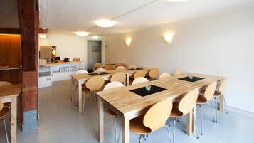 Un comedor con varias mesas y sillas de madera, una cocina al fondo, y un interior blanco con vigas de madera.