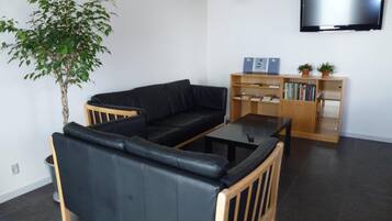 Ein Wohnzimmer mit einem schwarzen Ledersofa, einem Couchtisch aus Holz, einem Bücherregal, einer Topfpflanze und einem an der Wand befestigten Flachbildfernseher.