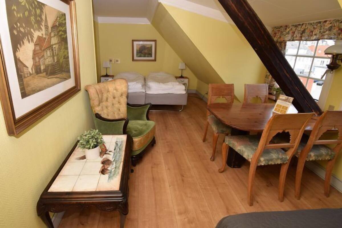 A cozy room with a bed, a dining table with chairs, a green armchair, and a small table with a plant.