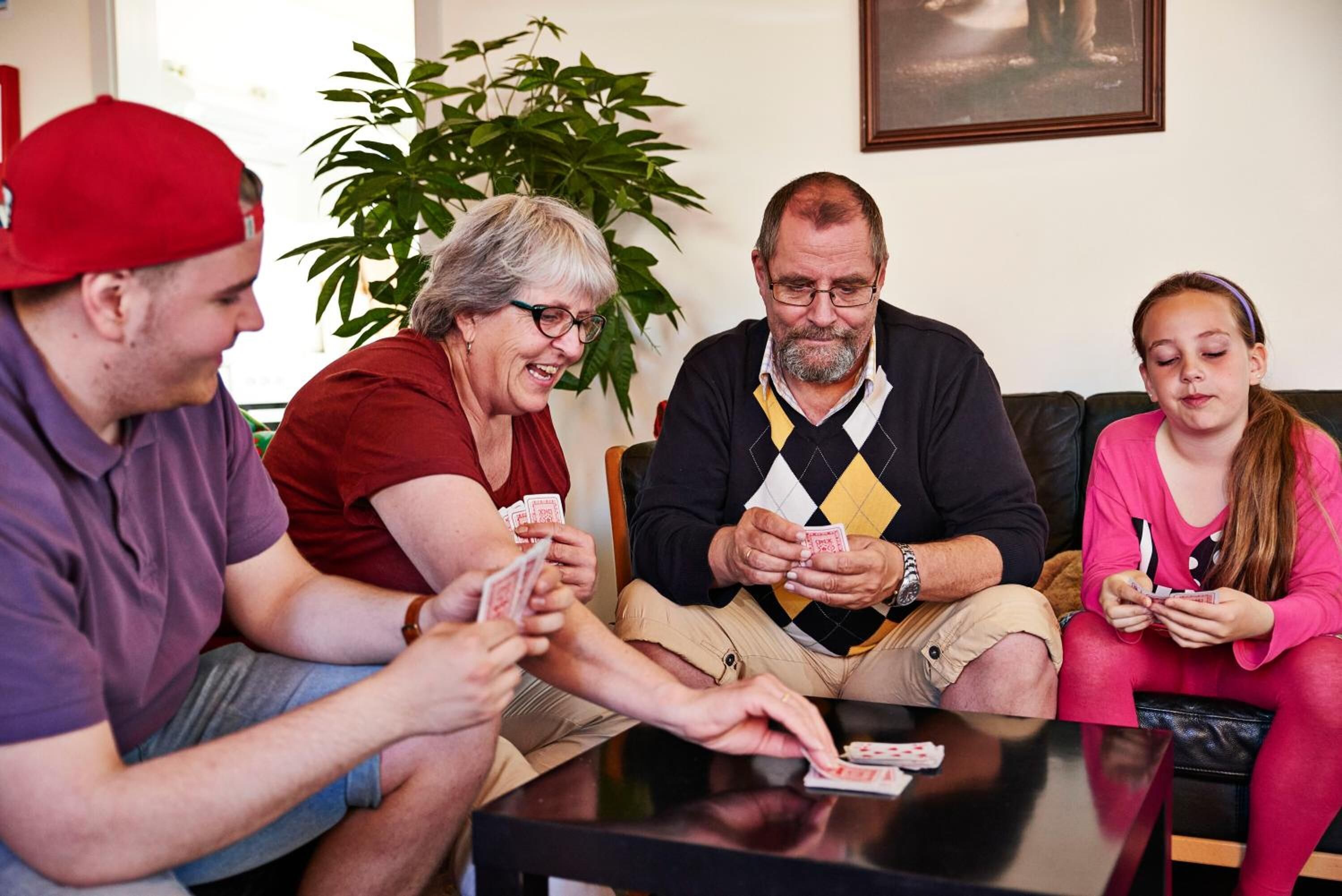 Four people playing cards at a table in a room with a plant and a framed picture on the wall.