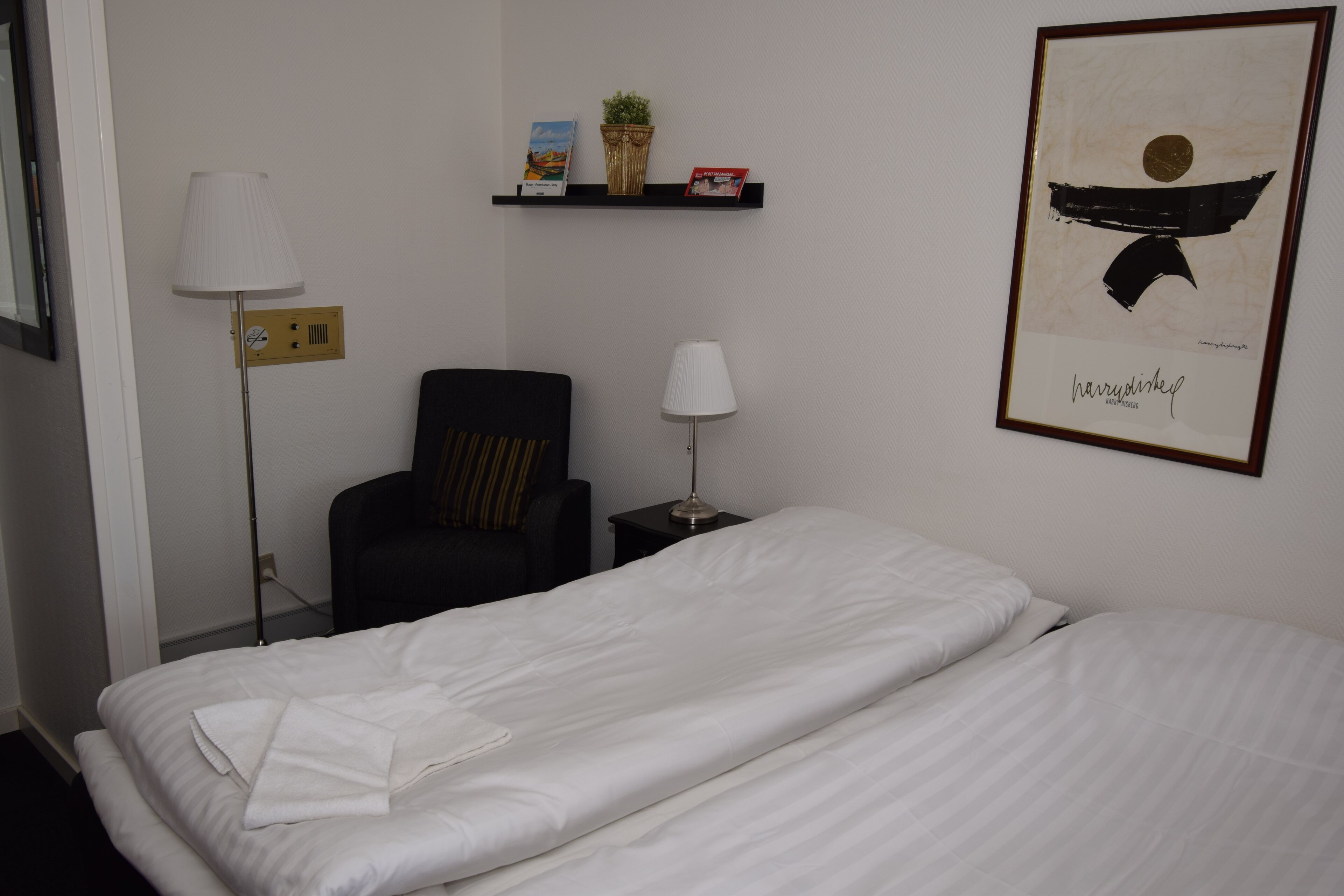 Habitación de hotel con cama, dos sillas, una mesita de noche, lámpara, estante con libros y una planta, y un cuadro en la pared.