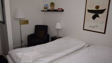Habitación de hotel con cama, dos sillas, una mesita de noche, lámpara, estante con libros y una planta, y un cuadro en la pared.
