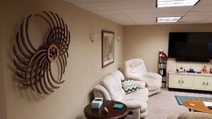 A living room with a beige sofa, a round coffee table, a decorative metal wall piece, a framed picture, and a TV on the wall. - Spacious suite with fenced back yard, deck, gas grill, fireplaces and splash pad (Athens)