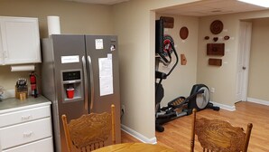 A compact kitchen with a refrigerator, a water dispenser, and a coffee machine. There are two chairs and a dining table. The room has a wooden floor and a door leading to another room with a treadmill. - Spacious suite with fenced back yard, deck, gas grill, fireplaces and splash pad (Athens)
