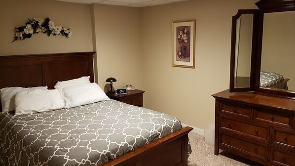 A neatly made bed with a patterned bedspread, a wooden nightstand with a lamp and clock, a dresser with a mirror, and a framed floral artwork on the wall. - Spacious suite with fenced back yard, deck, gas grill, fireplaces and splash pad (Athens)