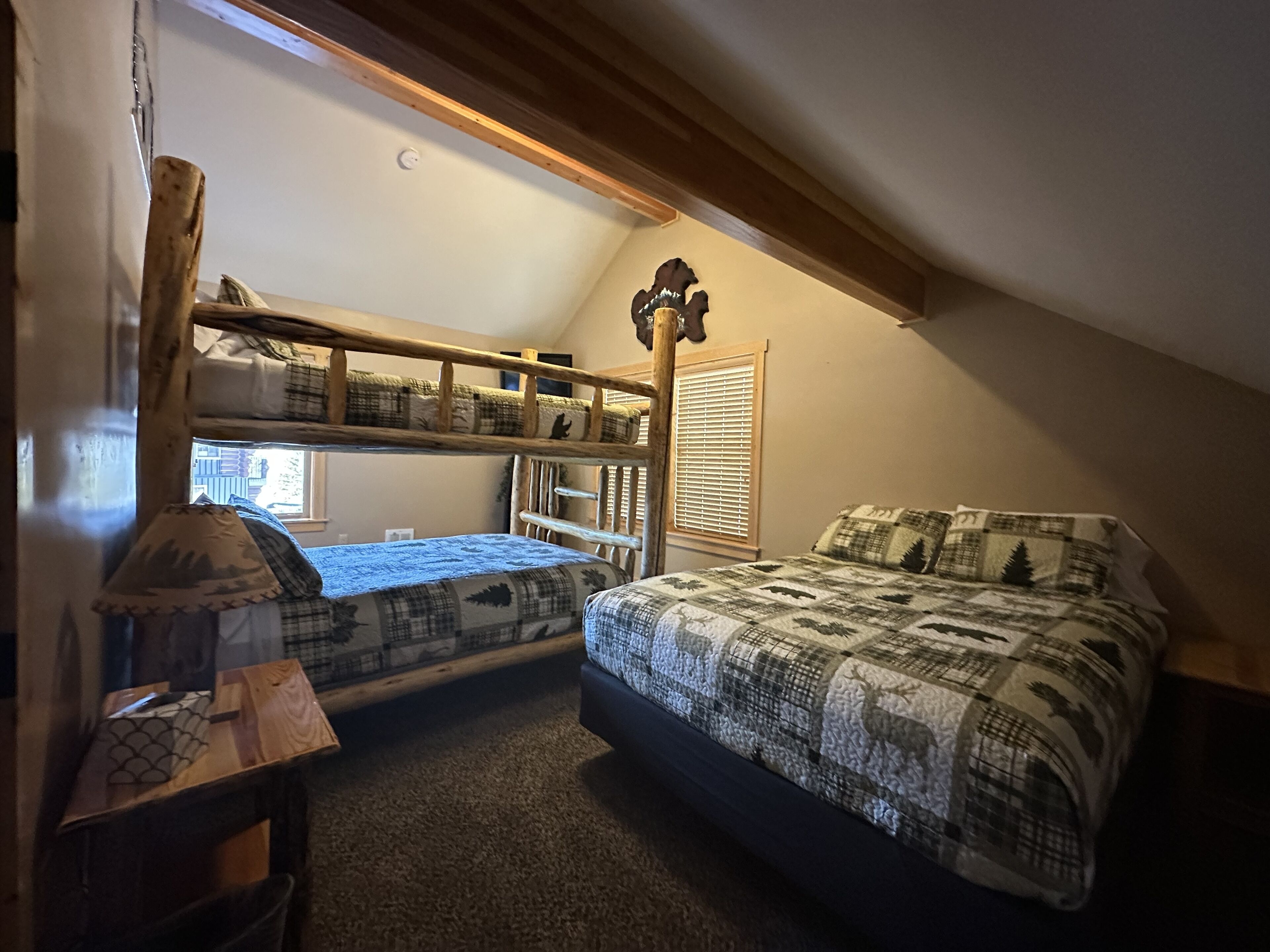 Ein gemütliches Schlafzimmer im Dachgeschoss mit Etagenbett und Einzelbett, Holzbalken, einem Fenster mit Jalousien und einem kleinen Tisch mit Lampe.