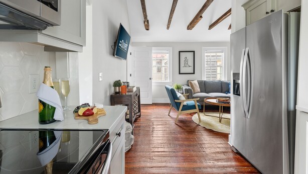 A modern kitchen with a stainless steel refrigerator, wooden beams on the ceiling, a dining area with a blue chair, and a living room with a gray sofa. - Casa Bella Suite B | 3 Blocks to King Street (Charleston)