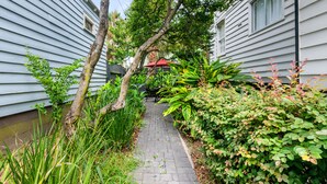 A narrow brick pathway flanked by lush greenery and two white buildings with windows. - Casa Bella Suite B | 3 Blocks to King Street (Charleston)