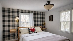 A bedroom with a checkered wall, a wooden bed, a bedside table with a lamp, and a window with blinds.
