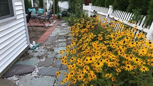 A garden with yellow flowers, a white fence, and a stone pathway leading to a patio area with outdoor furniture.