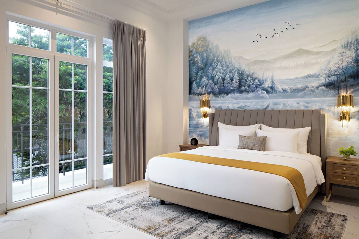 A bedroom with a large bed, a view of trees through the window, and a painting of a snowy landscape.