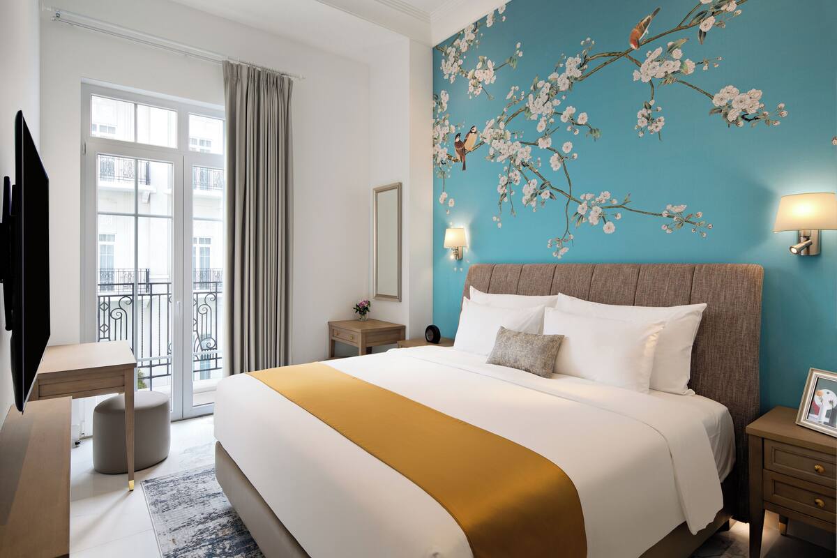 A hotel room with a large bed, a desk, a chair, a nightstand, a lamp, and a view of the outside balcony.