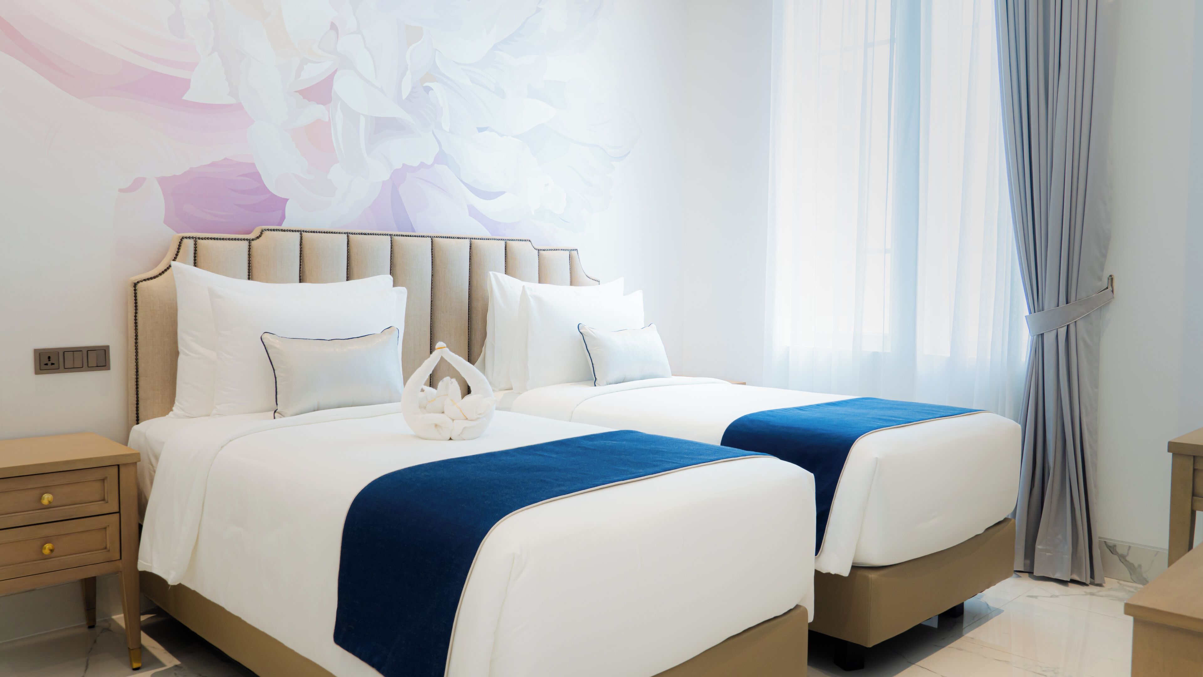 Cama doble con ropa de cama blanca y detalles azules, una mesita de noche y una pared con un mural floral.