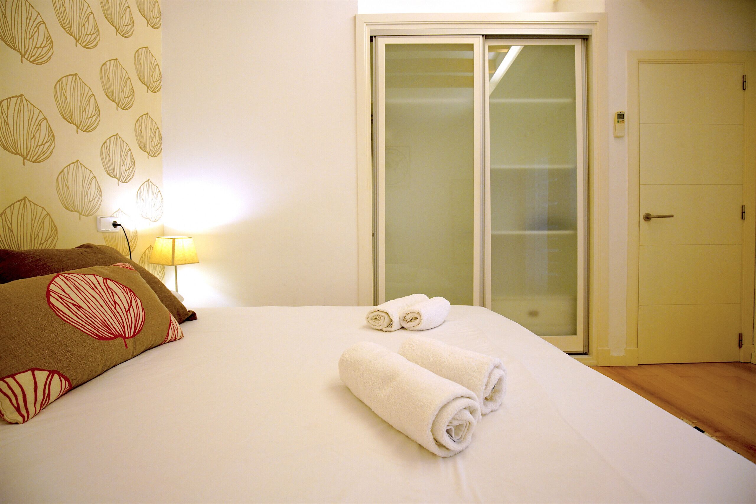 Una cama bien tendida con dos toallas enrolladas, una lámpara y una puerta con un panel de vidrio esmerilado.
