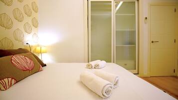 Una cama bien tendida con dos toallas enrolladas, una lámpara y una puerta con un panel de vidrio esmerilado.
