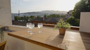 Een balkon met twee glazen witte wijn, een bloempot en een uitzicht op een stadsbeeld met gebouwen en bergen in de verte.
