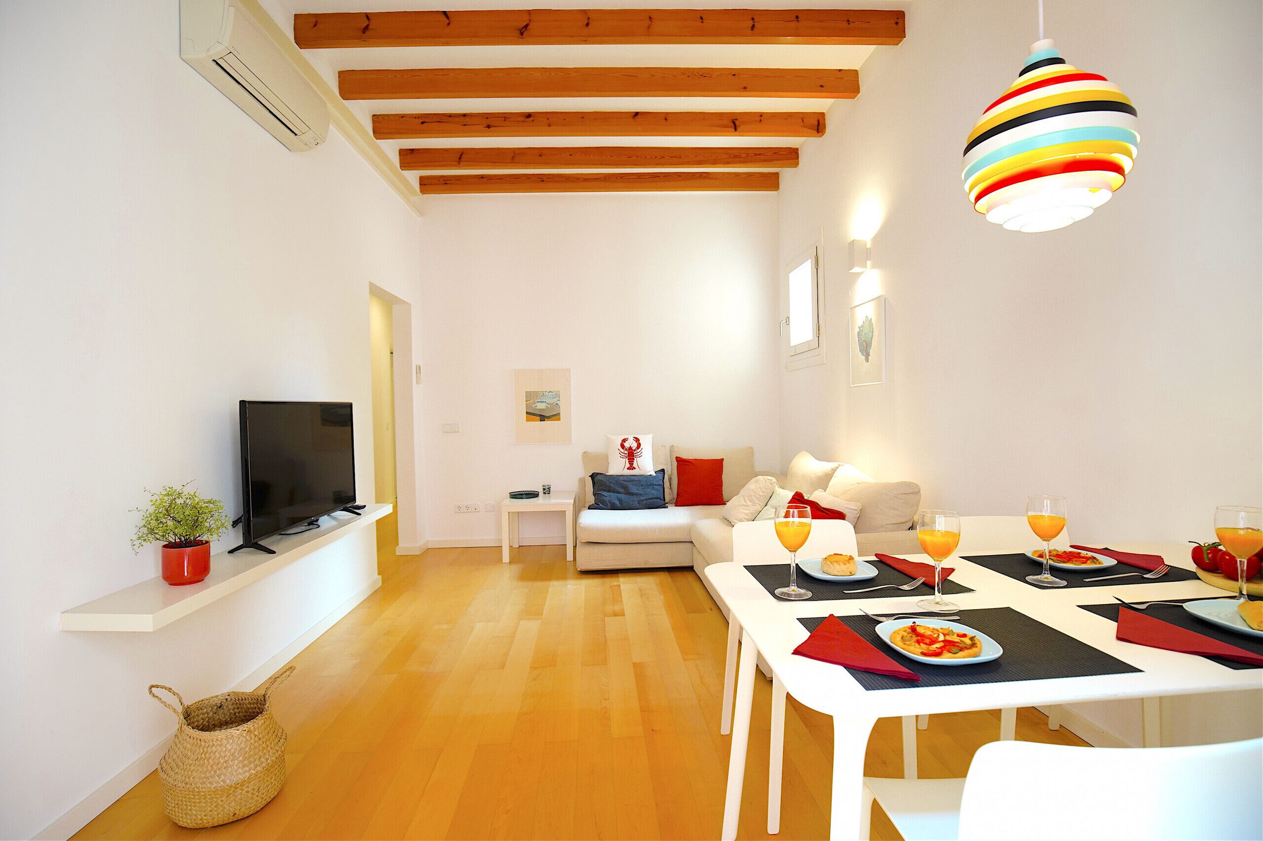 Een moderne woonkamer met een flatscreen-tv, een bank, een eettafel gedekt voor twee personen, compleet met sinaasappelsap, en een kleurrijke hanglamp.