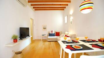 Een moderne woonkamer met een flatscreen-tv, een bank, een eettafel gedekt voor twee personen, compleet met sinaasappelsap, en een kleurrijke hanglamp.
