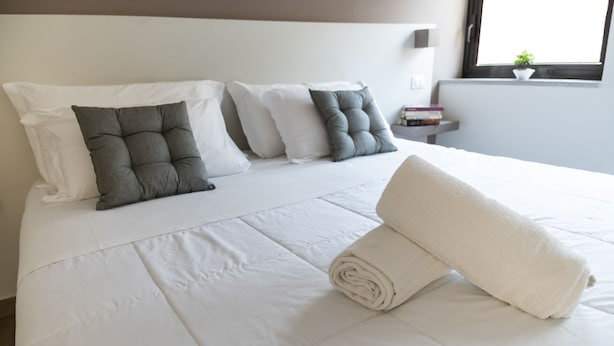 A neatly made bed with white bedding and a rolled towel, a bedside table with books, and a window with a plant.