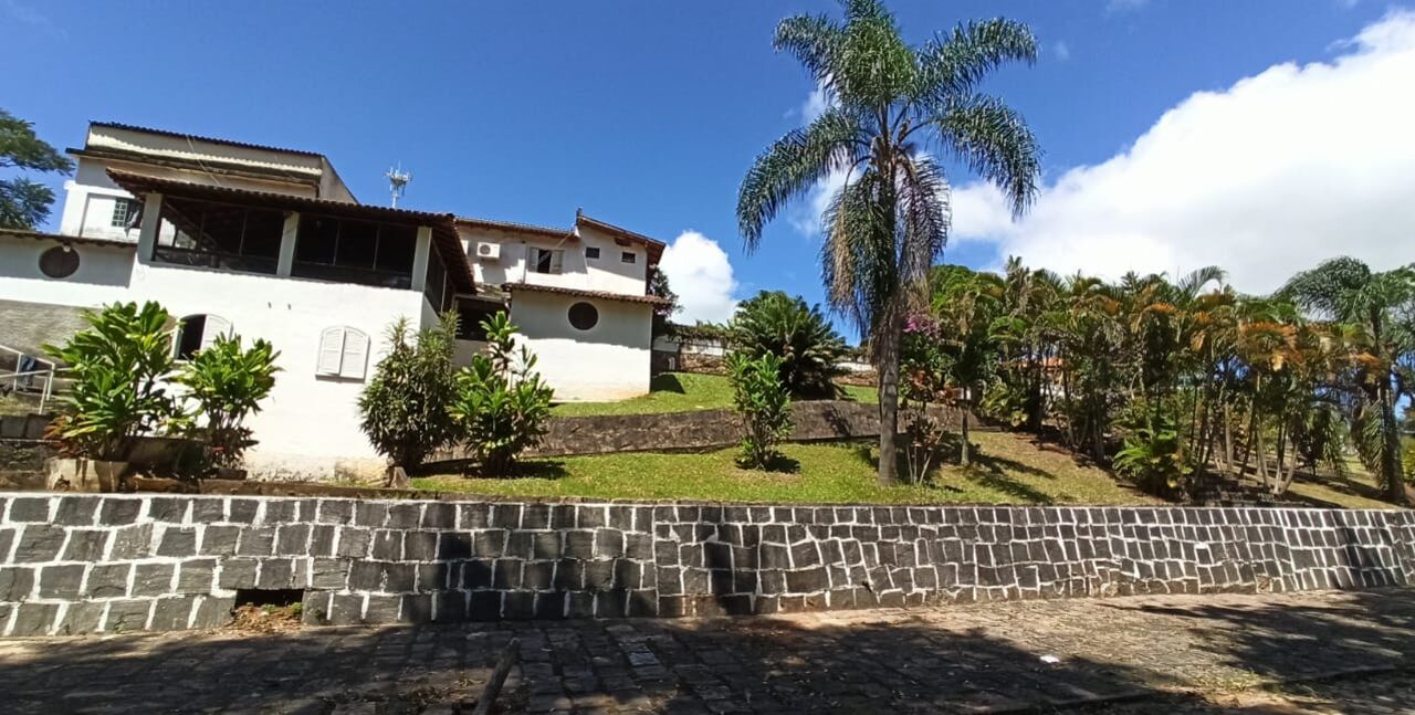 Um edifício branco com telhado ladrilhado, rodeado por um muro de pedra e vegetação exuberante, incluindo palmeiras.
