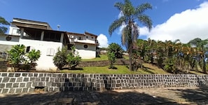 A white building with a tiled roof, surrounded by a stone wall and lush greenery including palm trees.