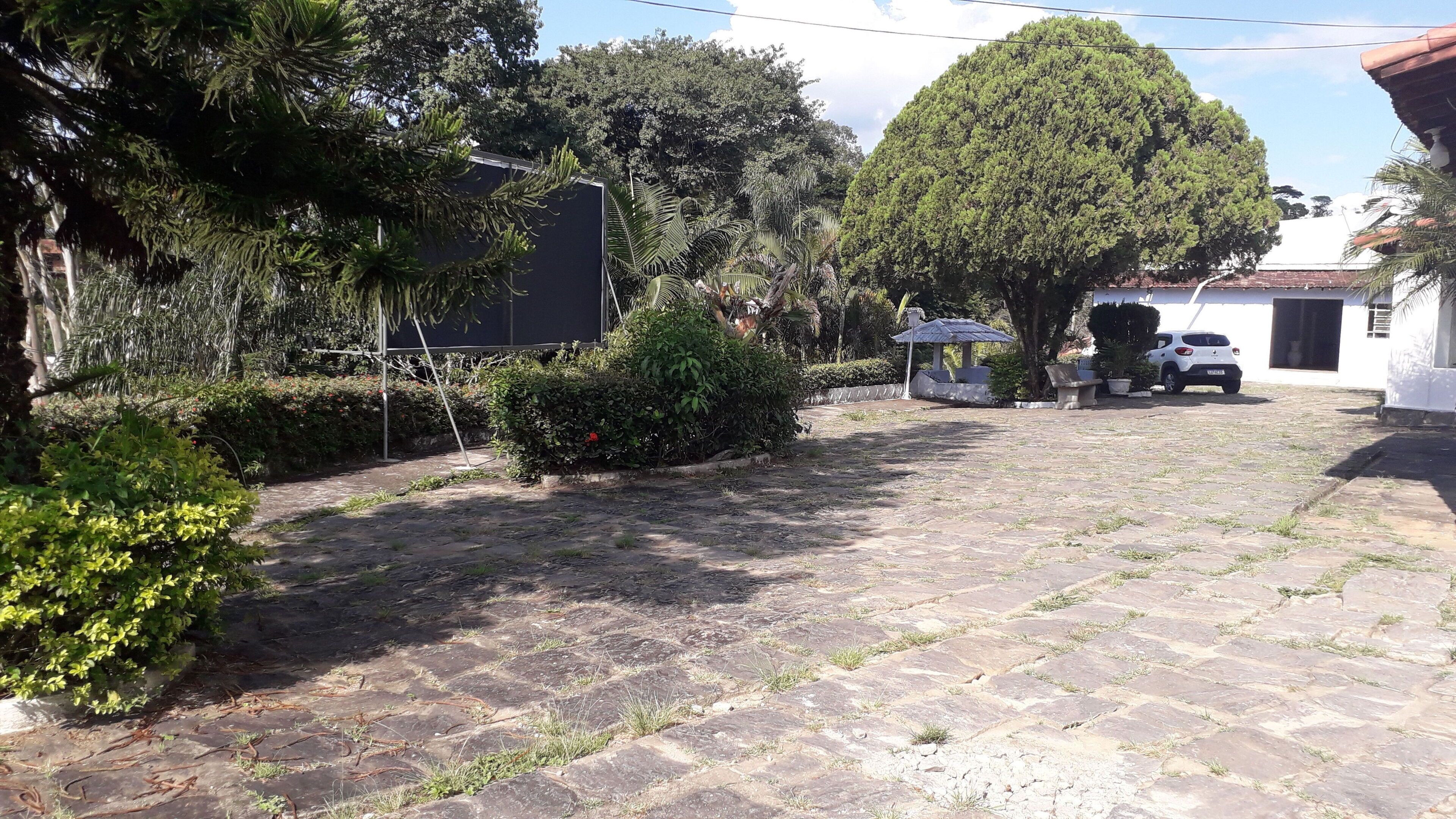 Un patio empedrado con baldosas de piedra, rodeado de abundante follaje y árboles. Un coche blanco está estacionado cerca de un edificio de fachada blanca.