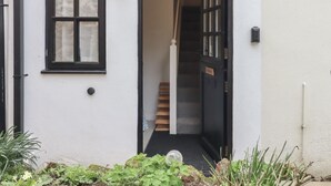 Un edificio blanco con un ventanal de marco negro y una puerta, un pequeño jardín con plantas, y una escalera que conduce a un piso superior.