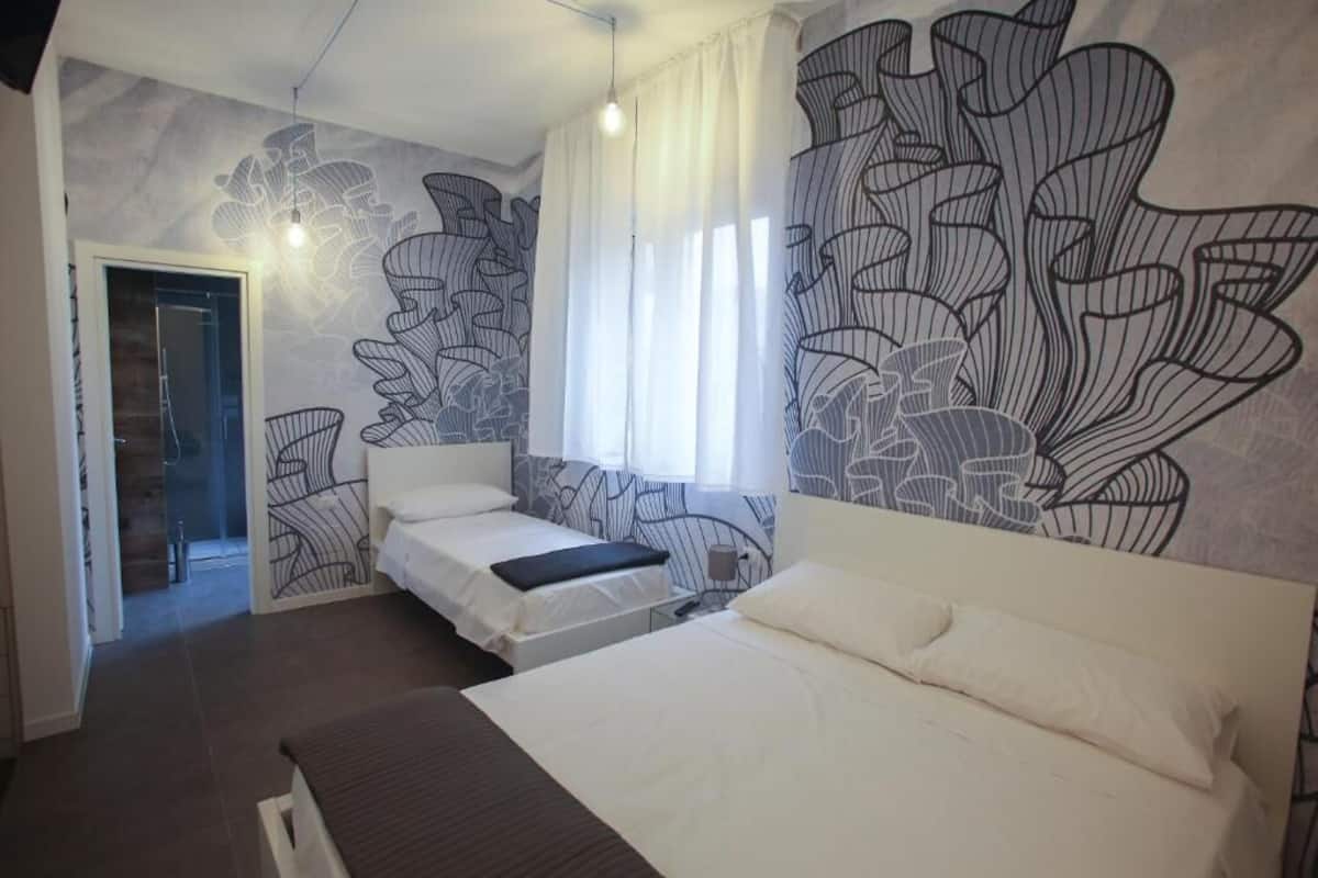 Ein Hotelzimmer mit zwei Betten, einem großen abstrakten Wandbild, einem Fenster mit weißen Vorhängen und einer Tür zu einem anderen Raum.