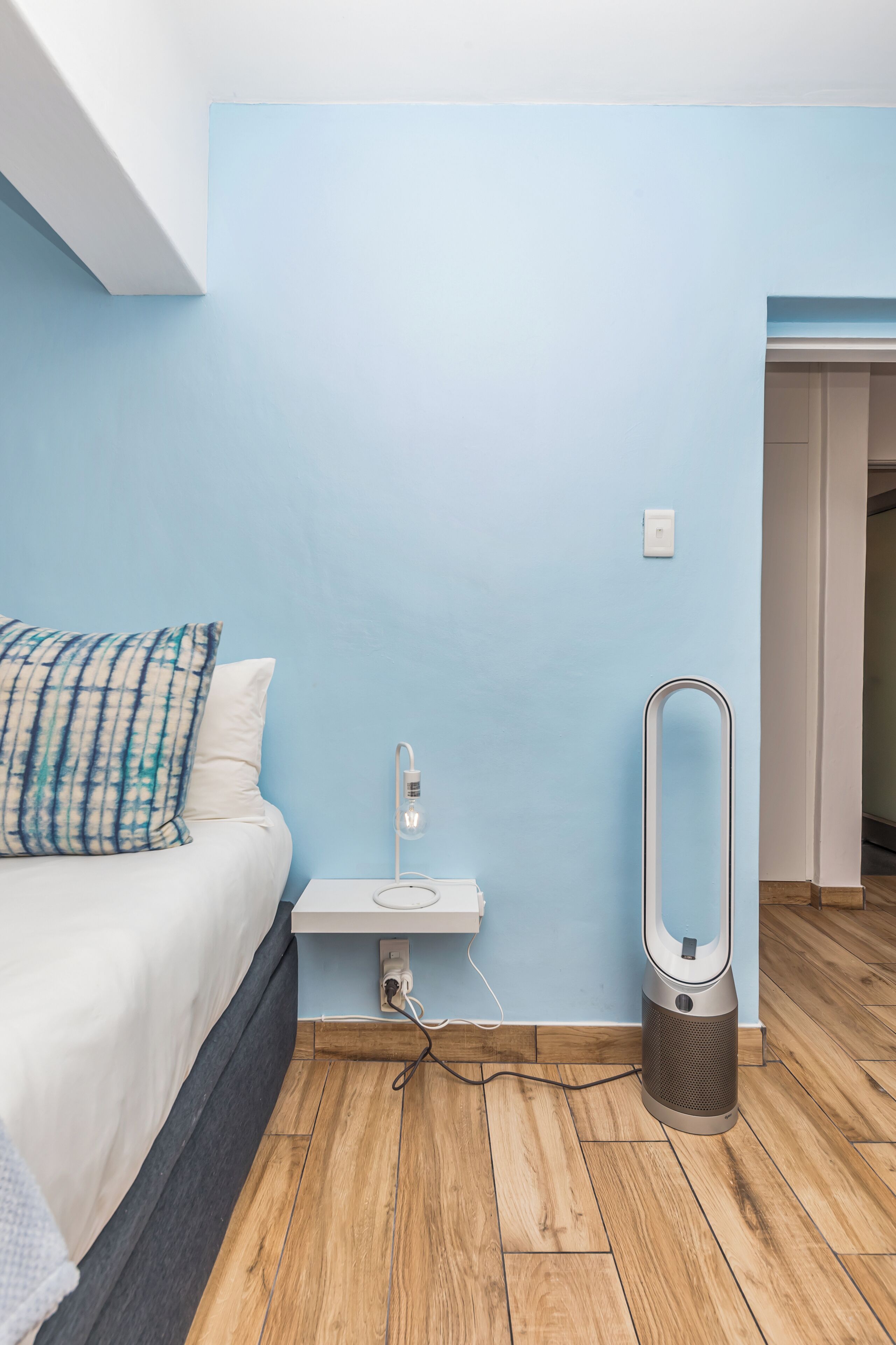 A bedroom with a bed, a bedside table, a standing heater, and a light switch on the wall.