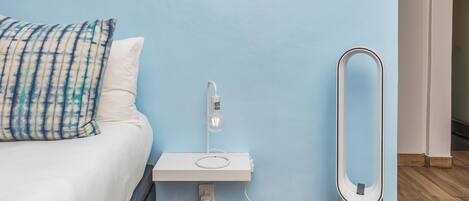 A bedroom with a bed, a bedside table, a standing heater, and a light switch on the wall.