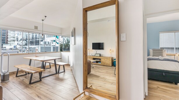 A modern room with a large mirror, wooden flooring, a bed, a TV, and a dining area with a table and benches. - Modern Space Footsteps from Sea Point Promenade (Cape Town)