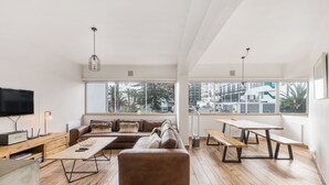 A modern living room with a sectional sofa, wooden coffee table, and a dining table with benches. Large windows offer a view of the city. - Modern Space Footsteps from Sea Point Promenade (Cape Town)