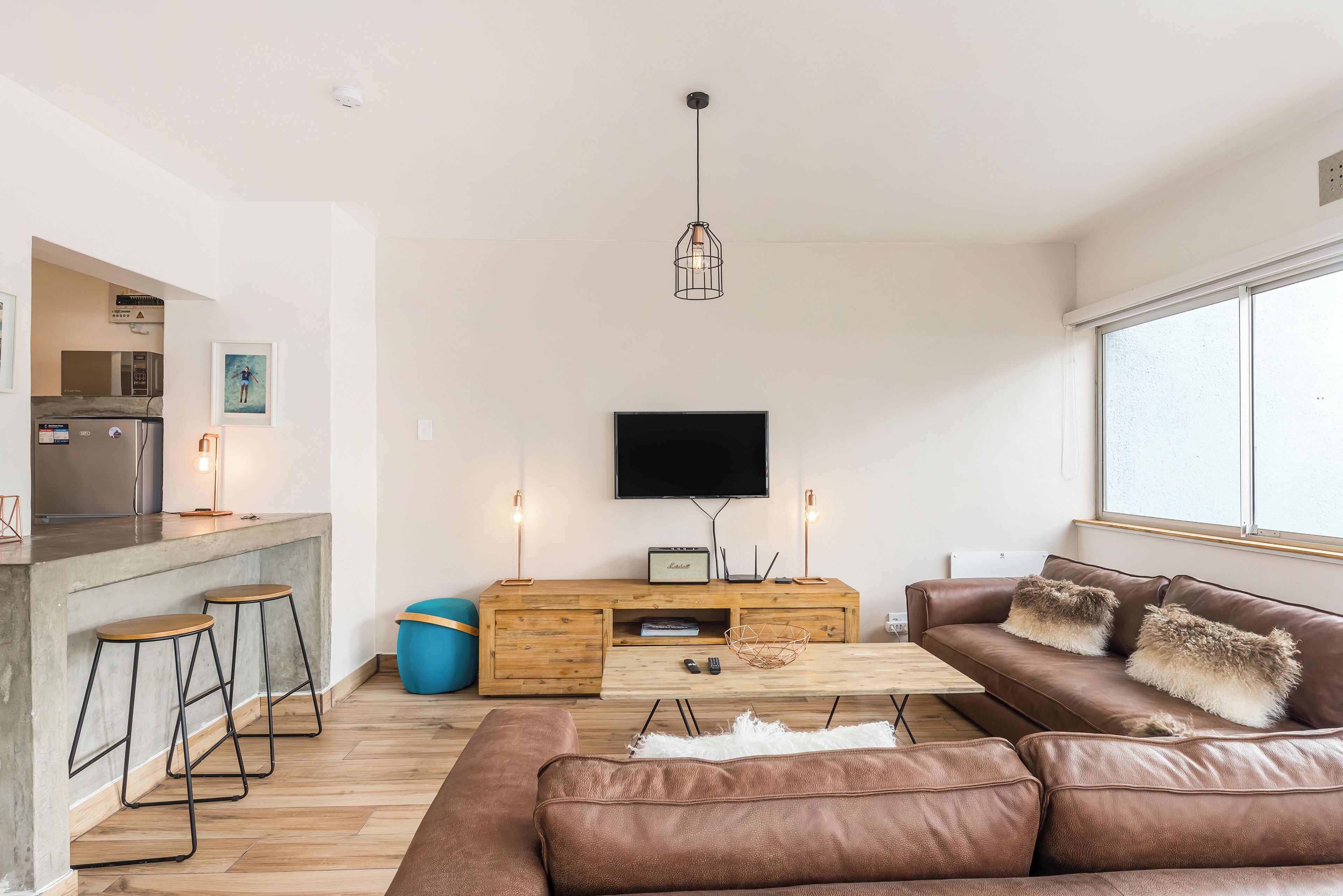 A modern living room with a brown leather sofa, a wooden coffee table, and a TV mounted on the wall. There are two bar stools and a blue ottoman.